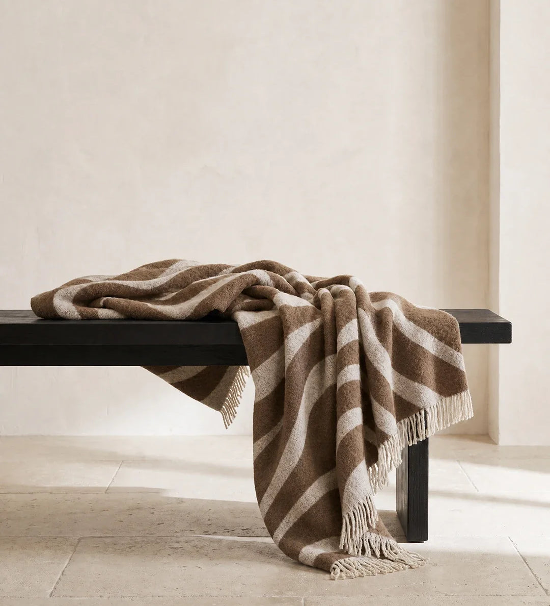 Pläd Flow Brun från Classic Collection i ull med vågformat mönster i brun och off-white, draperad över en mörk träbänk.