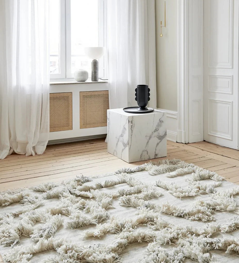 Melange, en handgjord ryamatta med hög struktur i ljusa naturtoner. Mattan ligger på ett ljust trägolv, omgiven av vita gardiner, marmorbord och dekorativa föremål i fönsternischen.