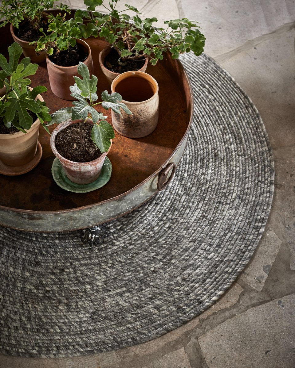 Round grey braided wool rug featuring a rustic metal tray holding assorted potted plants on a stone floor.