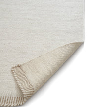Off-white woven wool area rug with a folded corner revealing a textured tan and cream striped border.