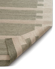 Vävd matta i ull med geometriskt mönster i gröna och beige nyanser, visad med ett uppvikt hörn för att visa texturen.