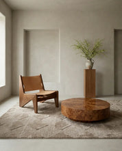 Modern tan leather lounge chair, round burl wood coffee table, and tall wooden pedestal on a geometric area rug.