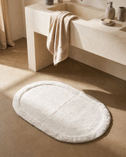 Vit oval badrumsmatta Border Oval från Classic Collection, 60x90 cm i bomull, placerad framför ett ljust handfat i ett varmt neutralt badrum.