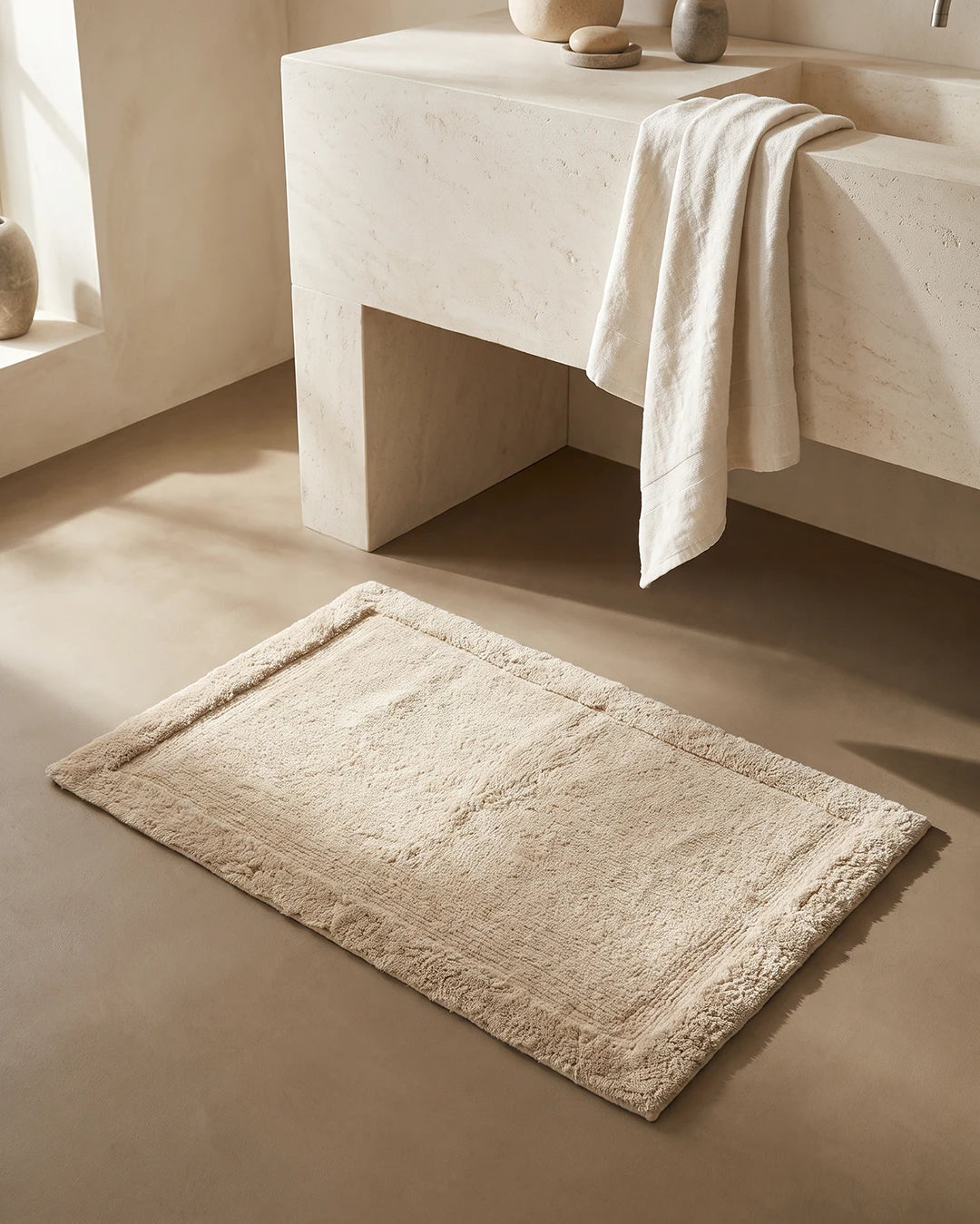 Badrumsmatta Border Beige från Classic Collection, 60x90 cm i bomull, placerad framför ett ljust handfat i ett varmt neutralt badrum.