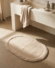 Badrumsmatta Border Oval Beige från Classic Collection, 60x90 cm i bomull, placerad framför ett ljust handfat i ett varmt neutralt badrum.