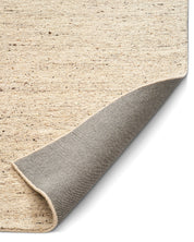 Närbild av Bouclé-matta i ljus beige ton med mörk melering och uppvikt hörn som visar den vävda baksidan.