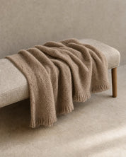 Pläd Bouclé Beige från Classic Collection i mjuk, strukturerad kvalitet med fransar, draperad över en ljus bänk.