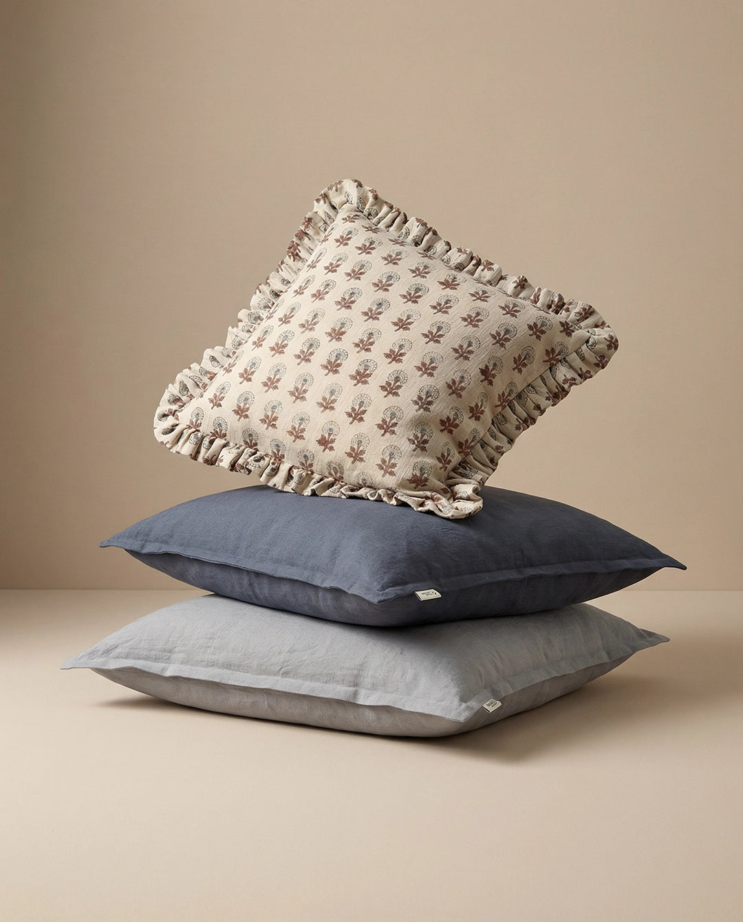 Stack of three linen throw pillows featuring a floral patterned ruffled cushion atop navy and grey solid square pillows.