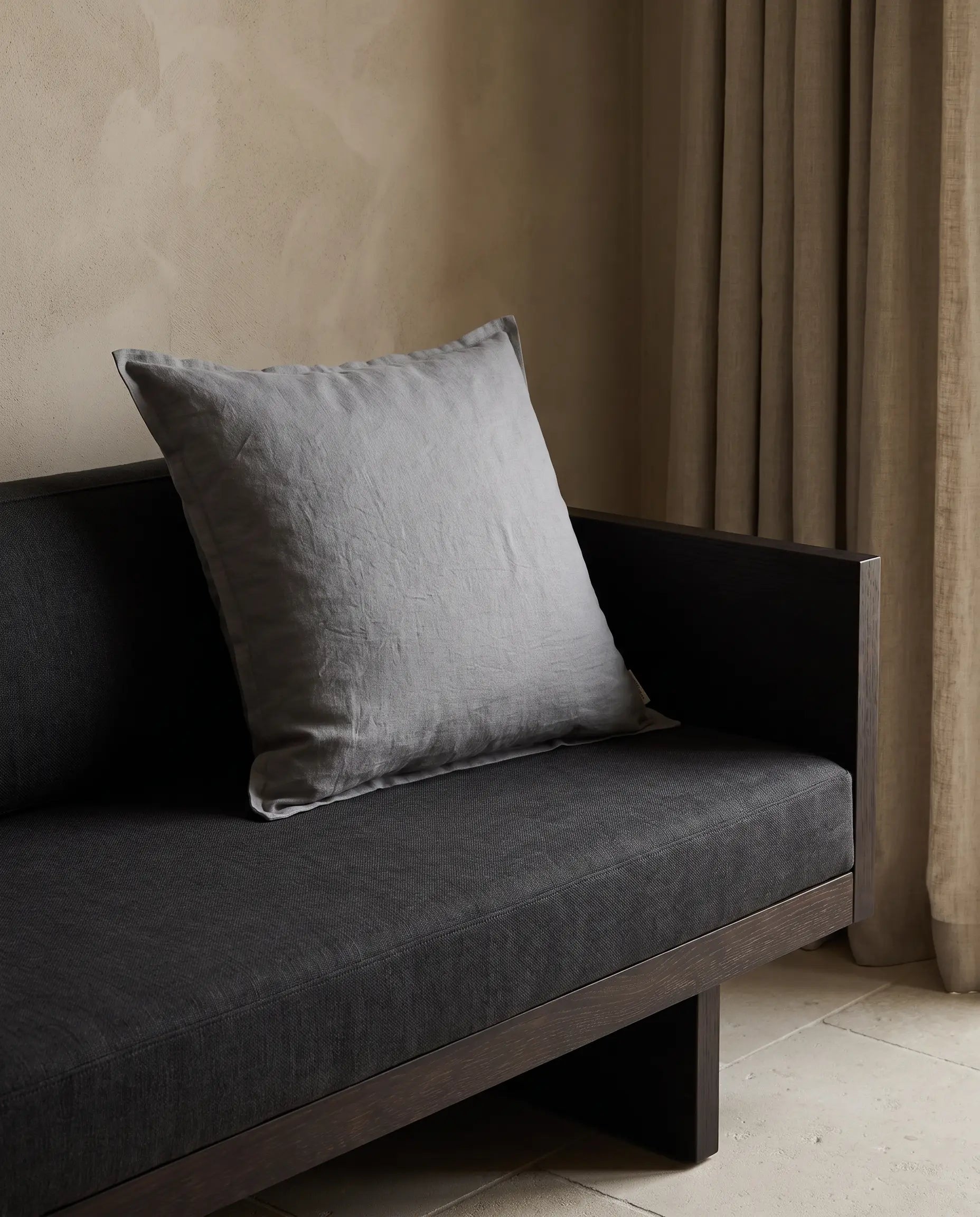 Kuddfodral Classic Linen Duvblå 50x50 cm från Classic Collection i enfärgat linne, stylat på en mörkgrå soffa i en varm, neutral interiör.