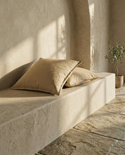Two square beige linen throw pillows placed on a textured stone bench in a warm, sunlit interior space.