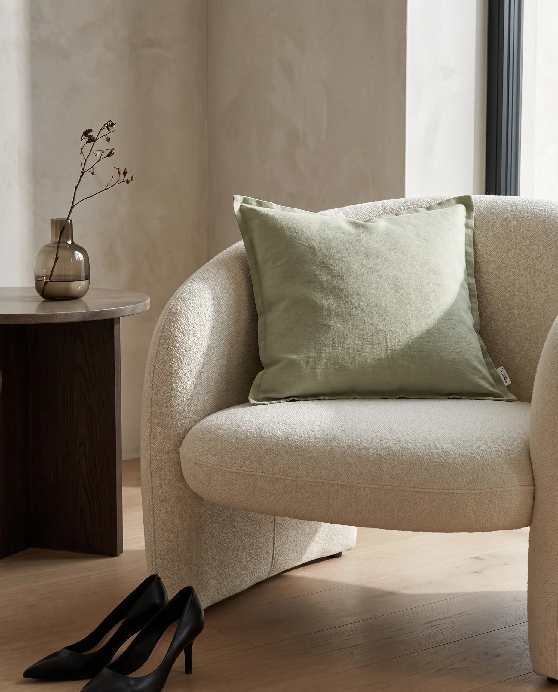 Kuddfodral Classic Linen Sage 50x50 cm från Classic Collection i enfärgat linne, stylat i en ljus fåtölj i en varm, neutral interiör.