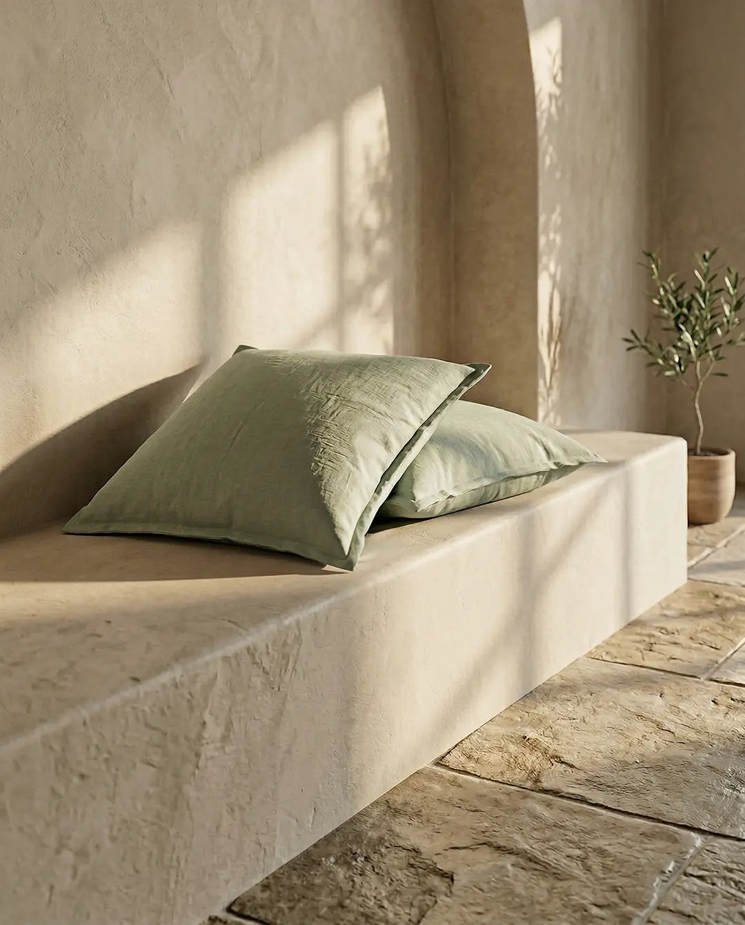 Två kvadratiska, salviagröna prydnadskuddar i linnelook placerade på en beige stenbänk i en rustik miljö.