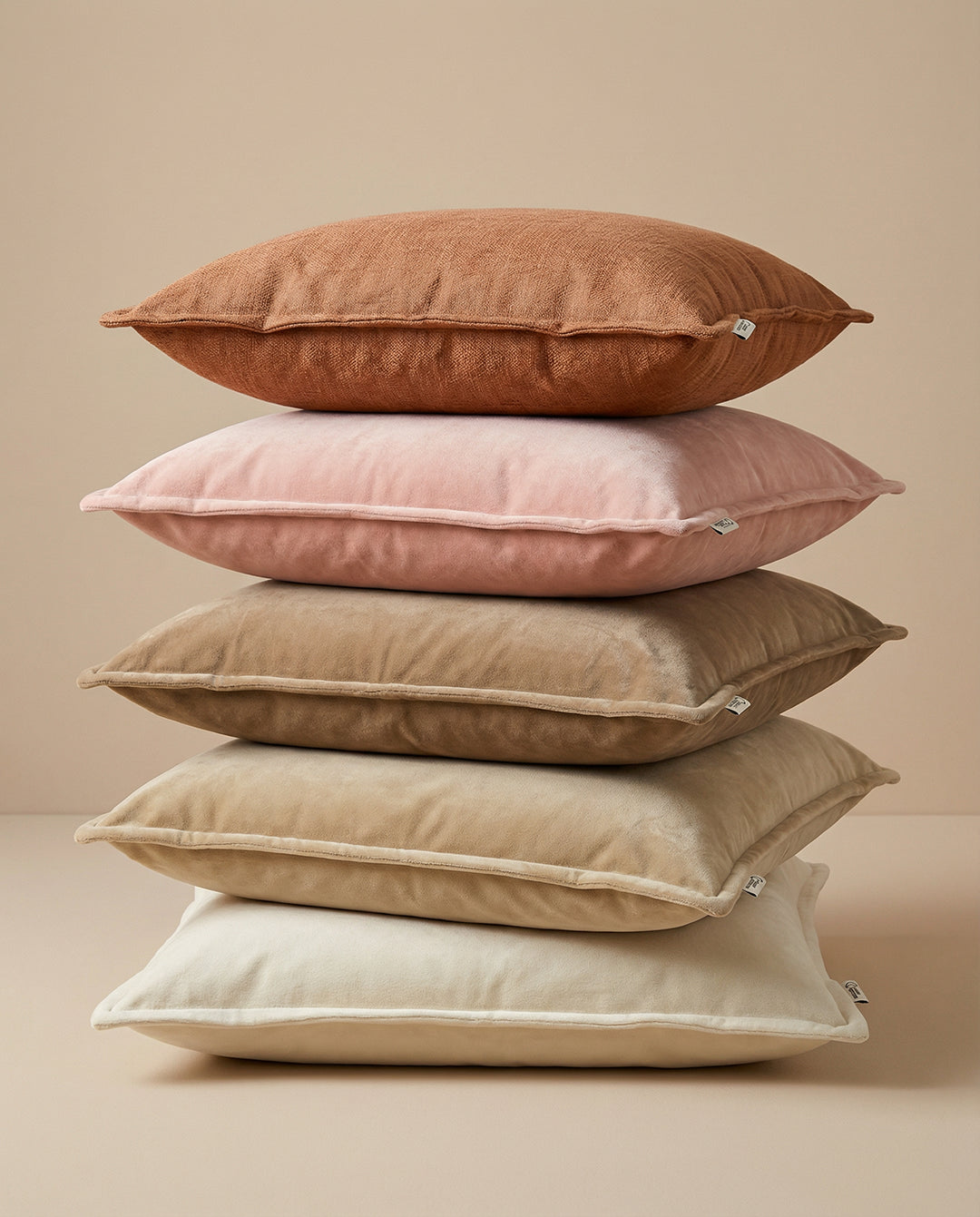 Stapel med fem kvadratiska prydnadskuddar i mjuka nyanser av rosa, beige och terrakotta i sammet och linne.