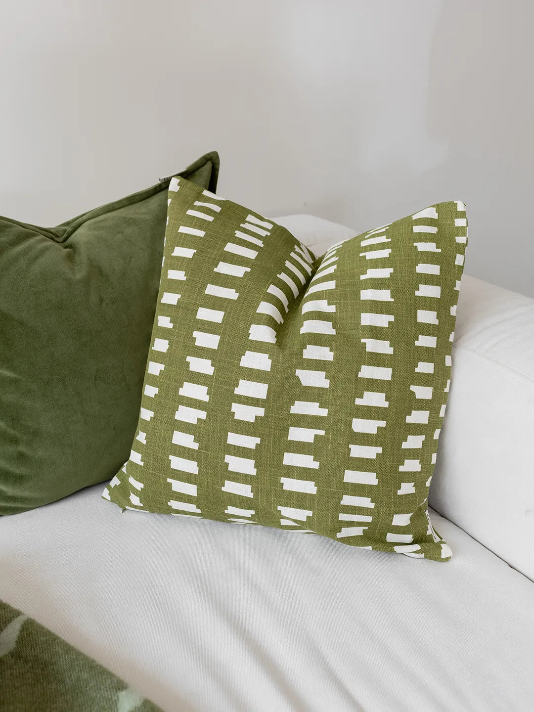 Kuddfodral Ikat Block Grön i bomull med grafiskt mönster, placerat på vit soffa tillsammans med grönt sammetfodral.