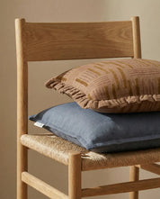 Två kuddar i linne med volangkant och geometriskt mönster placerade på en rustik trästol med flätad sits.