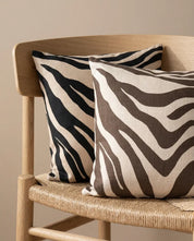 Two square throw pillows with beige and black/brown zebra print patterns on a woven wooden chair.