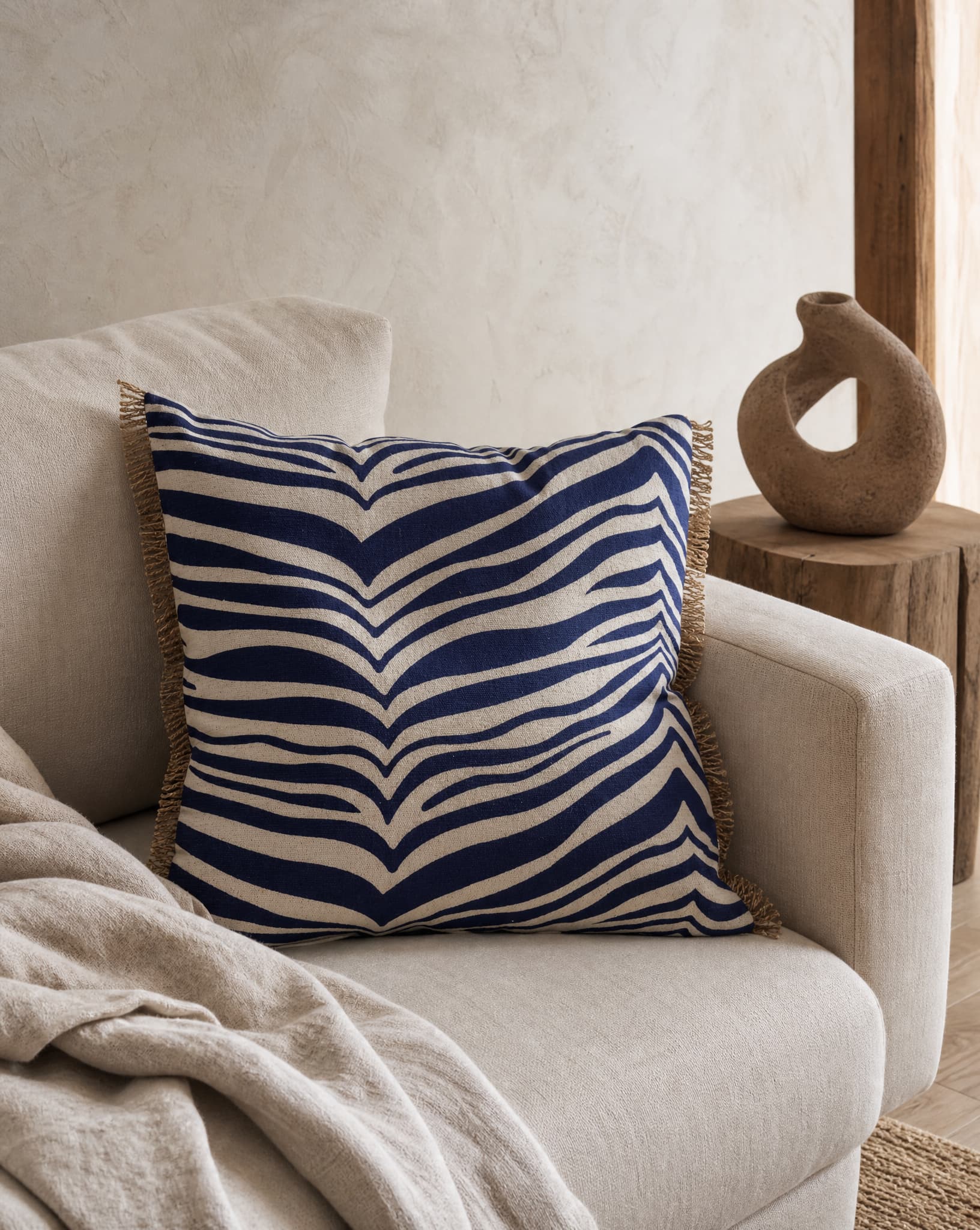 Kudde Zebra Navy, 50x50 cm, med grafiskt zebramönster i mörkblått och naturvitt samt dekorativa fransar längs sidorna, placerad i en ljus fåtölj i en varm och harmonisk inomhusmiljö.