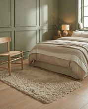 Beige shaggy area rug on light wood flooring in a cozy bedroom with sage green paneled walls and linen bedding.