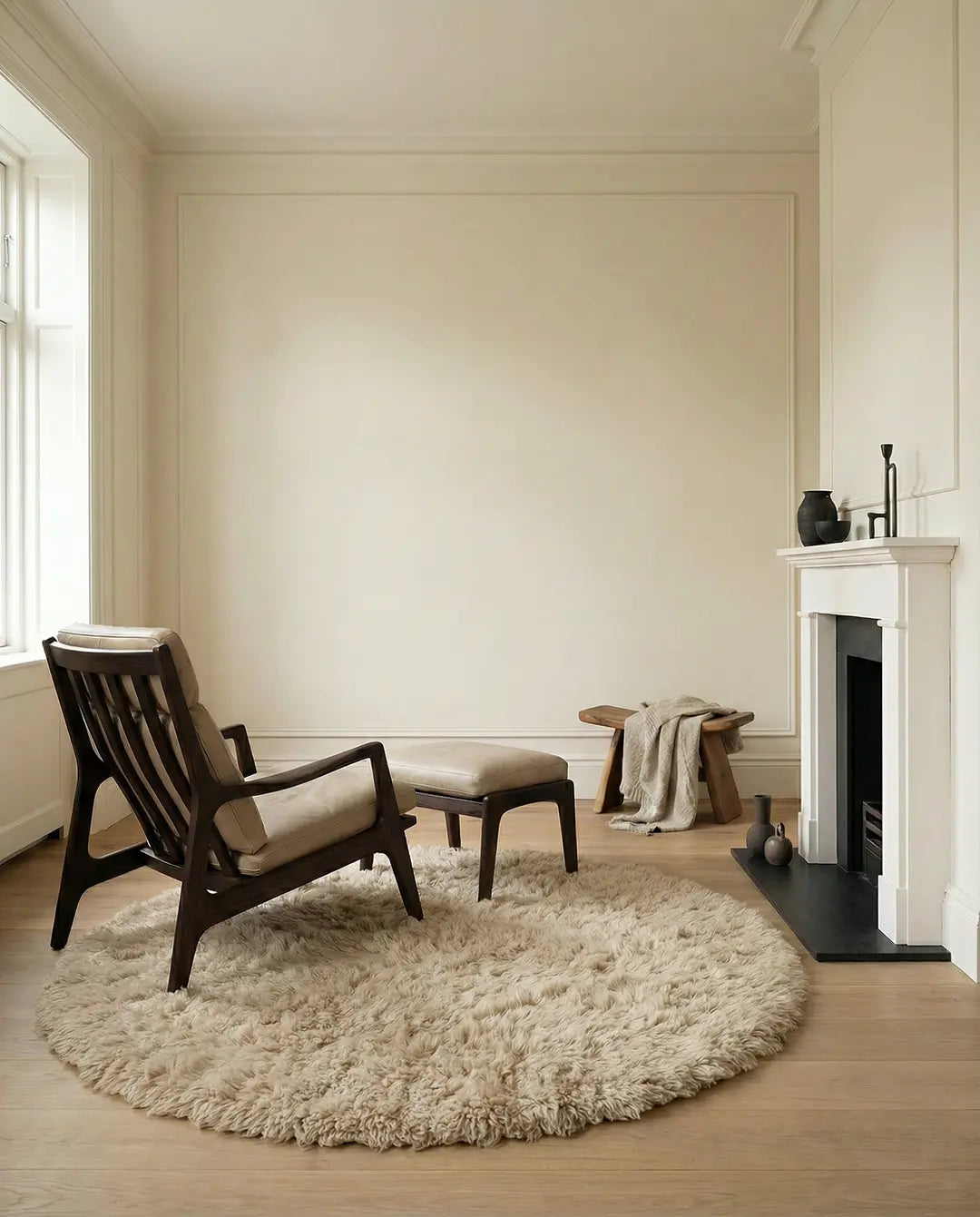 Mörk fåtölj i trä med beige läderdyna och matchande fotpall på en rund fluffig matta i ljust vardagsrum.