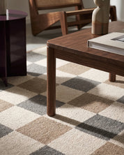 Närbild av handvävd ullmatta Check Grå/Natur från Classic Collection med rutmönster i grå, beige och naturvita toner, under ett mörkt träsoffbord intill ett mörkt vinrött sidobord.