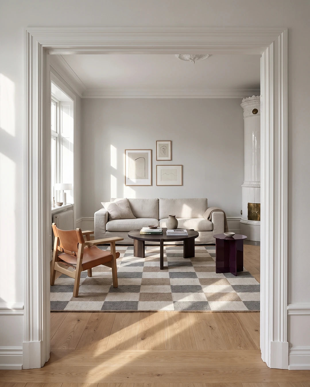 Handvävd ullmatta Check Grå/Natur från Classic Collection med rutmönster i grå, beige och naturvita toner, placerad i ett ljust vardagsrum med beige soffa, läderfåtölj, mörkt träsoffbord och vit kakelugn.