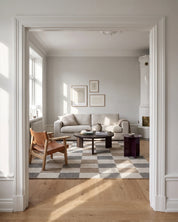 Handvävd ullmatta Check Grå/Natur från Classic Collection med rutmönster i grå, beige och naturvita toner, placerad i ett ljust vardagsrum med beige soffa, läderfåtölj, mörkt träsoffbord och vit kakelugn.