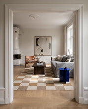Handvävd ullmatta Check Gul/Natur från Classic Collection med rutmönster i gult, beige och naturvitt, placerad i ett ljust vardagsrum med träsoffbord, grå soffa och fåtölj i trä.