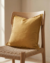 Mustard yellow square linen throw pillow resting on a wooden chair with a woven seat.