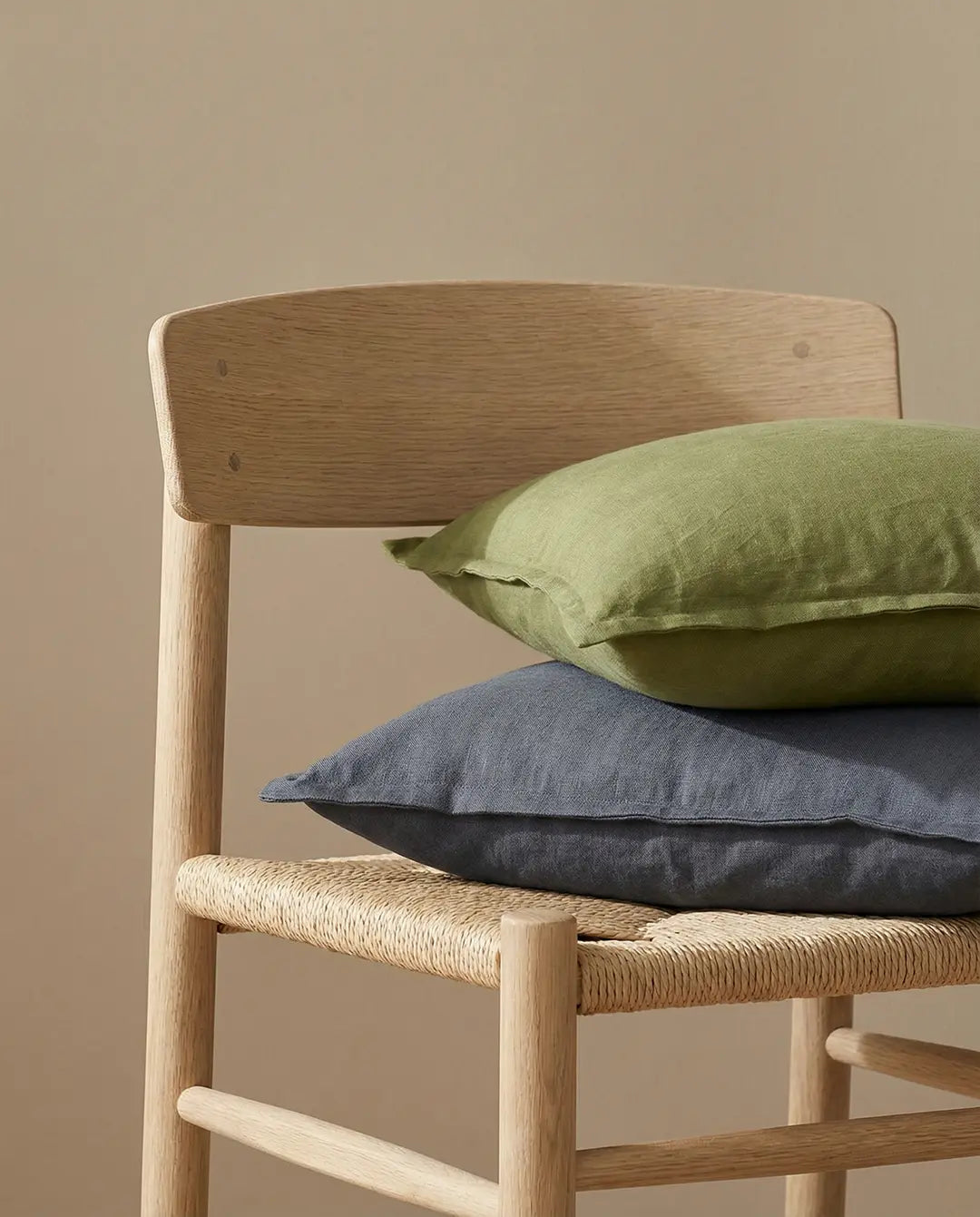 Two linen throw pillows in olive green and dark grey stacked on a light oak wooden chair with a woven seat.