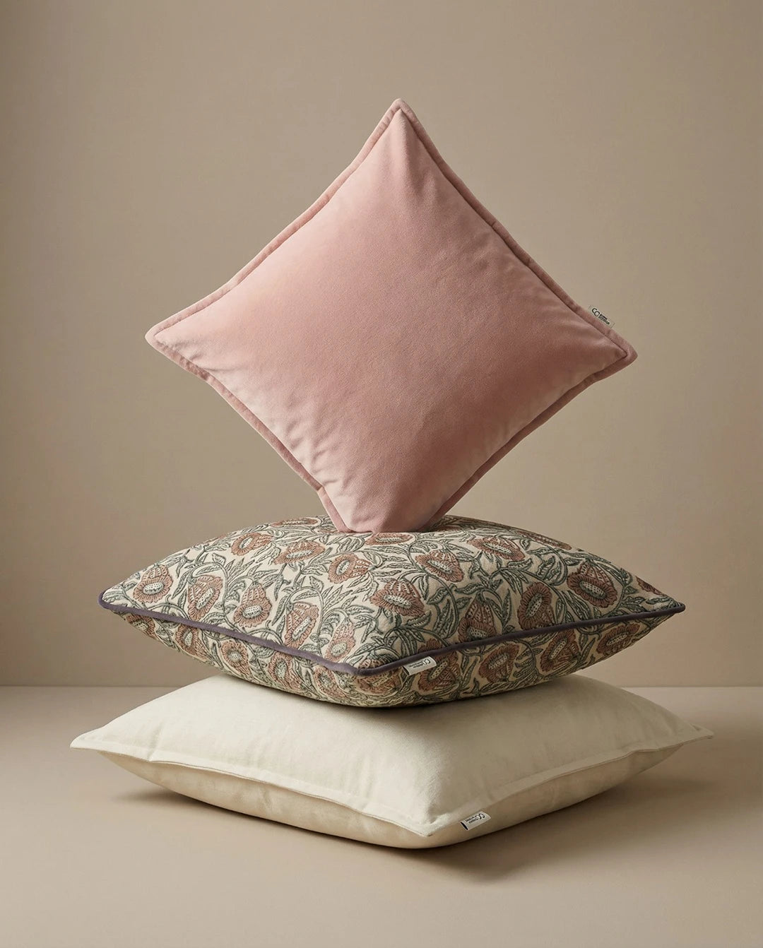 Tre staplade prydnadskuddar i rosa sammet, beige med blommönster och en ljusbeige kudde i en neutral miljö.