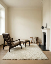 Dunkler Holz-Sessel mit beigem Lederpolster und Hocker auf einem flauschigen, cremefarbenen Teppich im modernen Wohnzimmer.