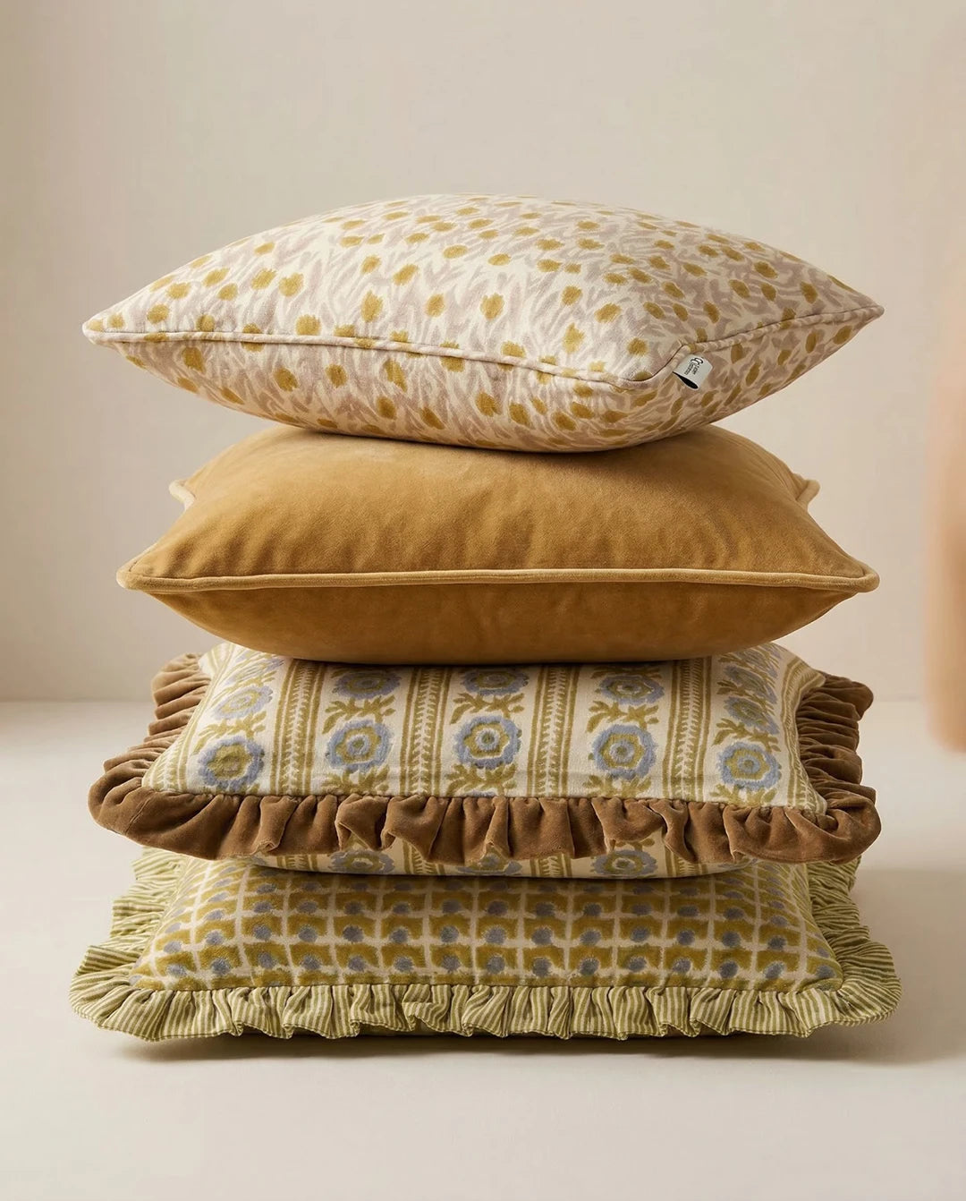 Stapel med dekorativa kuddar i olika nyanser av ockra, beige och blått med blommiga, rutiga och volangkantade detaljer.