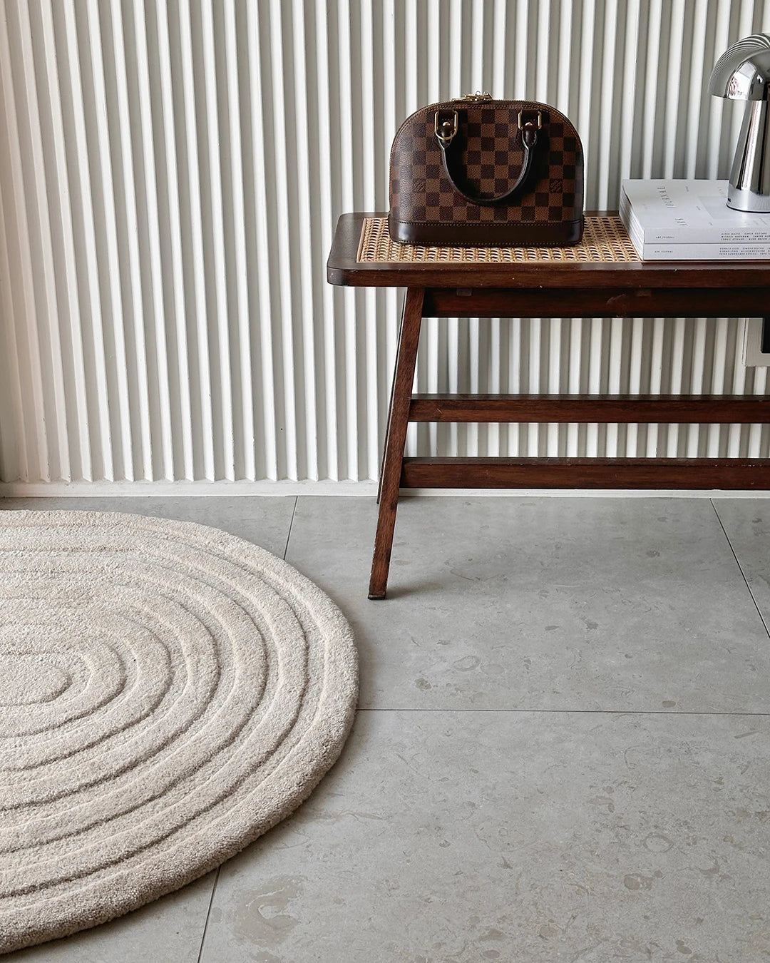 Handgjord dörrmatta Arch i färgen Beige, tillverkad i ull. Rund matta med mjukt skulpterat bågmönster placerad på stengolv i hallmiljö, intill träbänk och ribbad väggpanel.