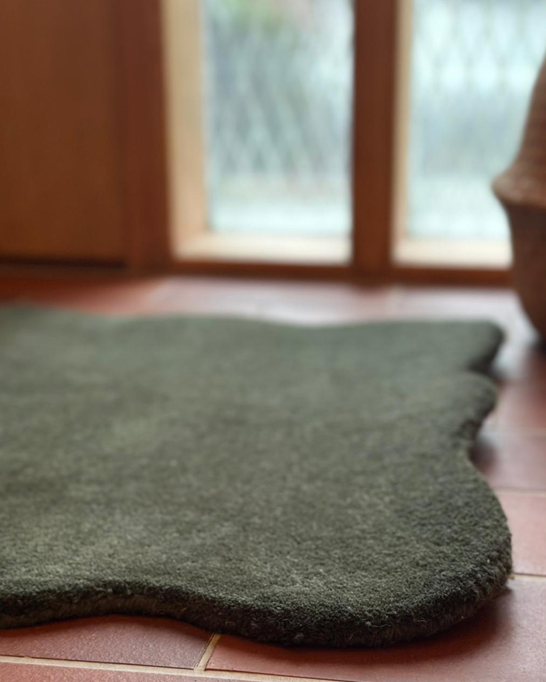 Dörrmatta Clam i färgen skogsgrön, 60x90 cm, tillverkad i ull med organiskt vågformad kant, fotograferad på rödbrunt klinkergolv vid fönster.