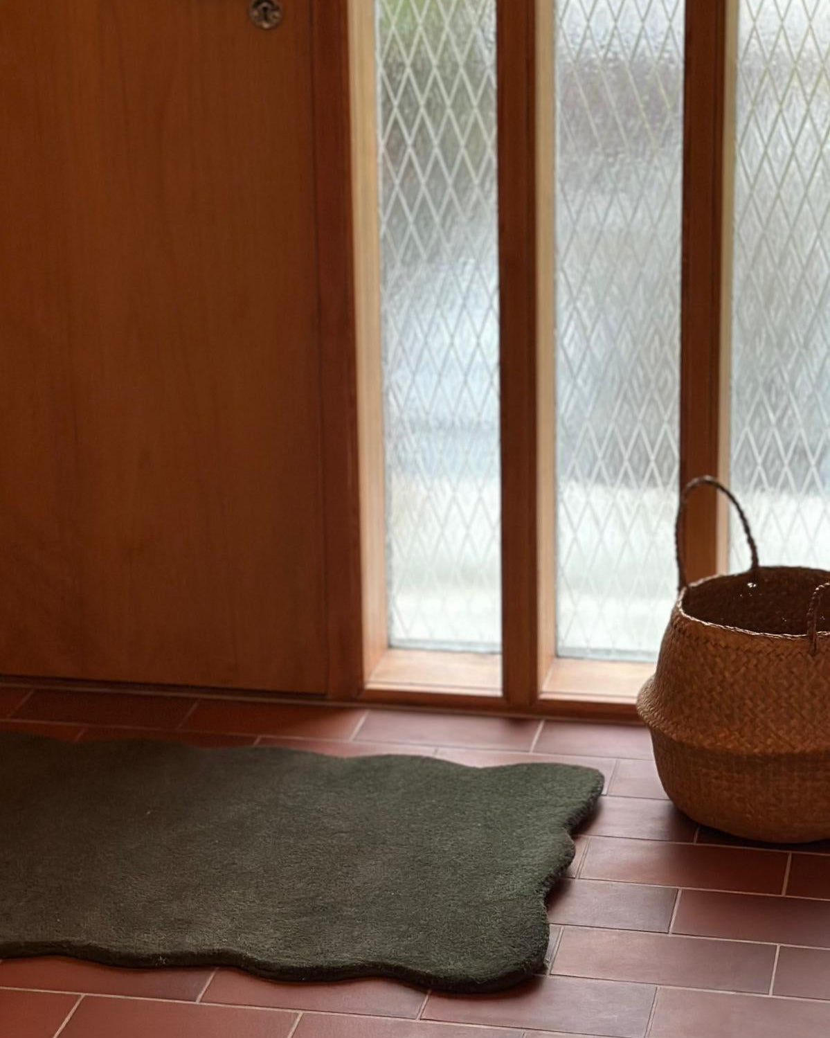 Dörrmatta Clam i färgen skogsgrön, 60x90 cm, tillverkad i ull med organiskt vågformad kant, fotograferad på rödbrunt klinkergolv vid dörr och fönster.