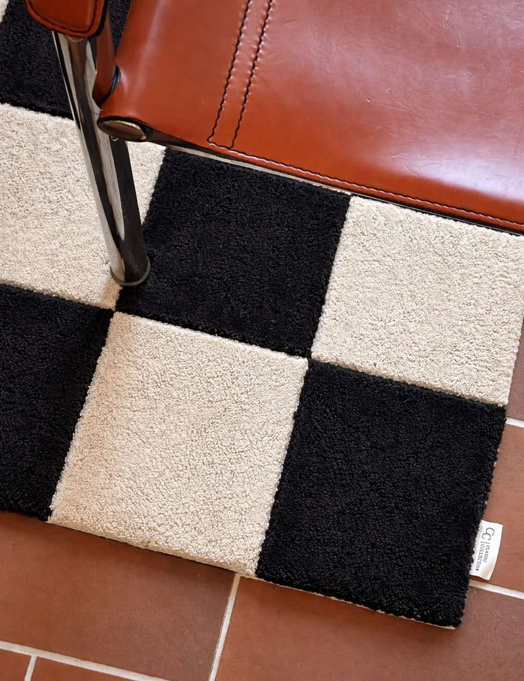 Dörrmatta Square i svart och naturfärgad ull med rutmönster, placerad i hallmiljö med stol och klinkergolv