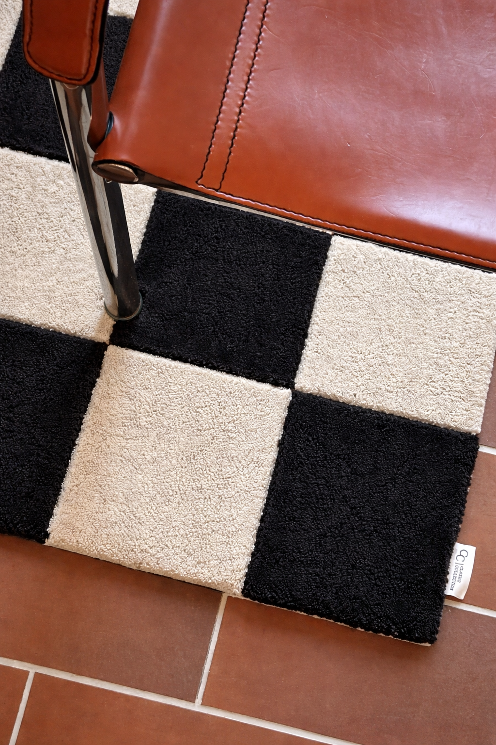 Dörrmatta Square i svart och naturfärgad ull med rutmönster, placerad i hallmiljö med stol och klinkergolv