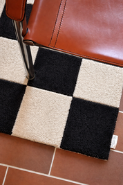 Dörrmatta Square i svart och naturfärgad ull med rutmönster, placerad i hallmiljö med stol och klinkergolv