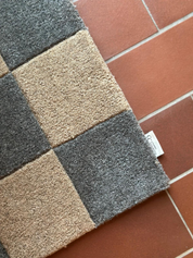 Dörrmatta Square i färgen grå/natur, tillverkad i ull med rutmönster i två nyanser, fotograferad på rödbrunt klinkergolv.