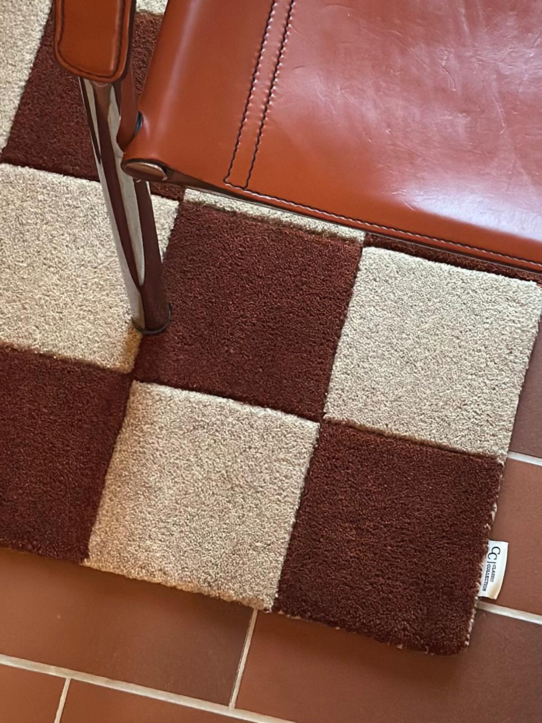 natur, tillverkad i ull med rutmönster i två nyanser, fotograferad på rödbrunt klinkergolv med brun läderstol i bild.