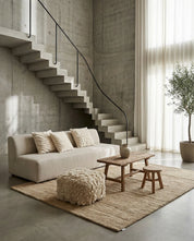 Beige linen sofa with textured throw pillows, a rustic wooden coffee table, and a shaggy ottoman in a modern concrete room.