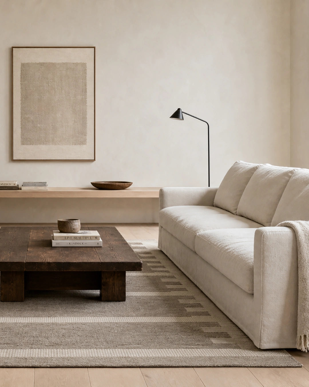 Ljust och minimalistiskt vardagsrum med beige soffa, mörkt soffbord i trä, två läderfåtöljer och en mönstrad ullmatta i neutrala toner. På väggen hänger ett abstrakt konstverk.