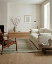 Beige soffa i ett minimalistiskt vardagsrum med trästolar och en grönrandig matta i naturlig stil.