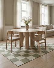 Runder Esstisch aus hellem Travertin mit Säulenfüßen, umgeben von Holzstühlen auf einem grün-weißen Schachbrett-Teppich.