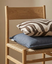Stacked brown zebra print and solid blue linen throw pillows resting on a light oak wood chair with woven seat.