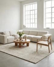 Modern cream sectional sofa with a round wooden coffee table and light wood chair on a textured grid-pattern area rug.