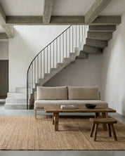 Beiges Sofa vor einer Betontreppe mit schwarzem Geländer, kombiniert mit einem rustikalen Holztisch und Juteteppich.