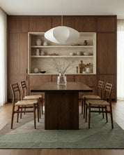 Modern walnut dining set with four woven-seat chairs and a minimalist pendant light in a wood-paneled dining room.