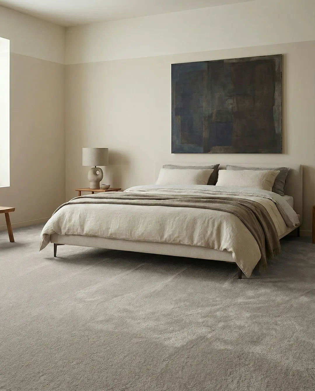 Minimalist bedroom with a beige upholstered bed, layered linen bedding, and a large abstract painting on a neutral wall.