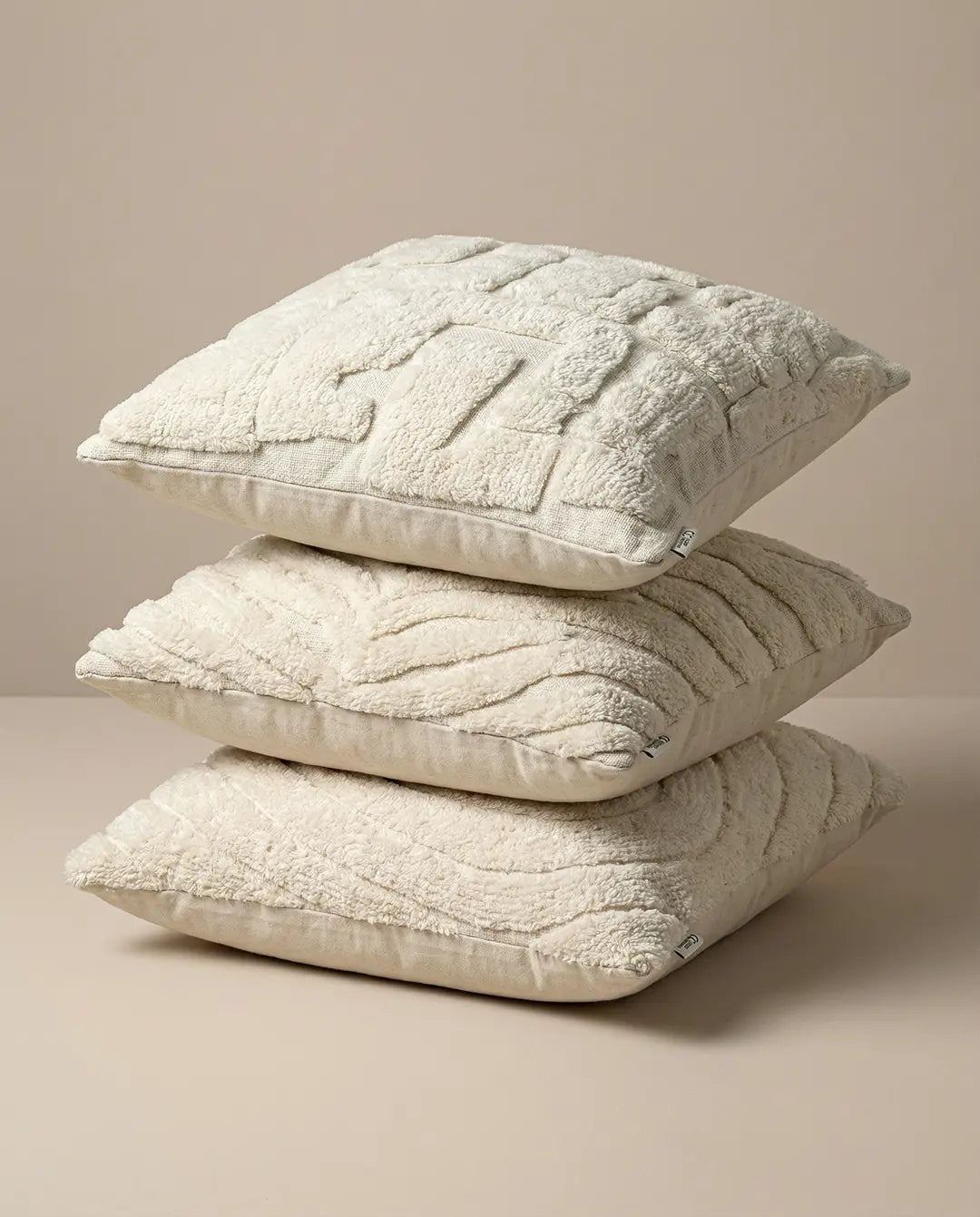 Stack of three square cream-colored throw pillows with textured, tufted patterns on a beige background.